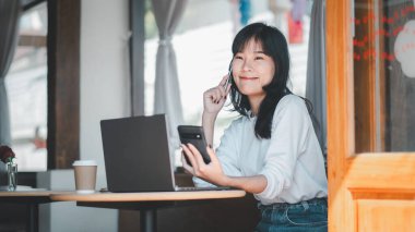 İş analitik kavramı, mutlu bir Asyalı kadın, bilgisayarını parlak ve dost canlısı bir kafe ortamında kullanırken akıllı telefonuyla hoş bir konuşma yapar.