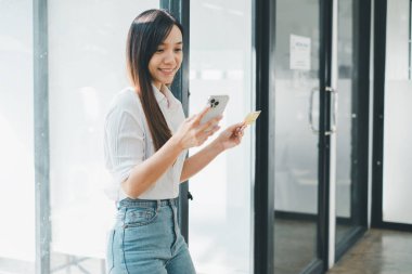 Çevrimiçi ödeme kavramı, elinde kredi kartı olan mutlu genç bir kadın akıllı telefonunu açık ve modern bir ortamda kullanıyor..