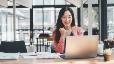 İş analitiği konsepti, genç bir kadın, parlak, modern bir kafede bilgisayarının üzerinde çalışırken başarılı bir anı kutlarken heyecan ve neşe gösteriyor.