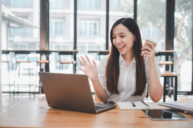 İş analizi konsepti, gülümseyen uzaktan kumandalı bir işçi dizüstü bilgisayarına el sallıyor, iyi aydınlatılmış bir ofiste elinde kahveyle bir video konferans görüşmesi yapıyor.