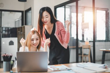 İş analitiği konsepti, iki neşeli Asyalı iş kadını modern ofis ortamında başarılı bir projeyi kutluyor, bir dizüstü bilgisayar ve belgeler masanın üzerinde.