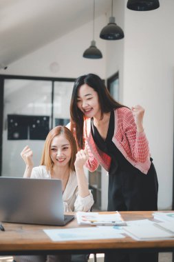 İş analitiği konsepti, iki memnun iş kadını çalışma masasında ortak bir başarıyı kutluyor, biri oturuyor, diğeri ayakta, ikisi de zafer işaretleri gösteriyor..