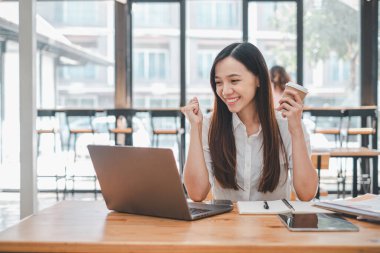 İş analitiği konsepti, gülümseyen genç bir kadın elinde kahve ve mutlu bir ifadeyle kafeterya masasında dizüstü bilgisayarla çalışmaktan zevk alır..