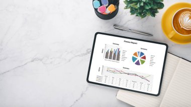 Renkli finansal grafikleri ve grafikleri gösteren dijital bir tablet, mermer bir yüzey üzerinde bir fincan kahve ve defterin yanında.