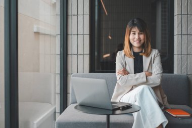 Kendine güvenen bir girişimci, modern bir kafede çapraz kollarıyla otururken gülümser. Bilgisayarı ve akıllı telefonu iş için hazırdır..