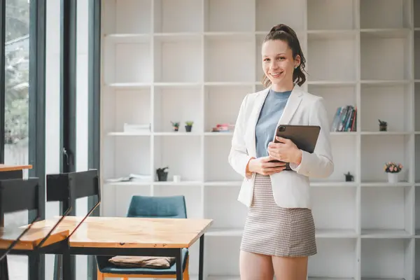 Smiling professional woman holding a tablet stands in a bright, contemporary office space with bookshelves in the background.