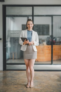 İyi organize edilmiş modern ofis ortamında elinde dijital tablet tutan şık beyaz ceketli gülümseyen profesyonel bir kadın..