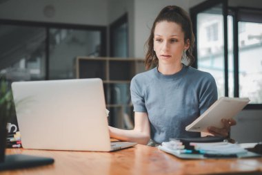 Çalışkan bir kadın notlarını özenli bir gözle gözden geçirir. Laptop ekranındaki verilerle karşılaştırır. İyi aydınlatılmış, modern bir çalışma alanında..