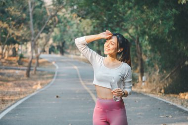 Işıl ışıl genç bir kadın alnından teri siliyor, ferahlatıcı bir egzersizden sonra gülümsüyor, bileğinde akıllı bir bantla bir su şişesini tutuyor doğal bir park ortamının ortasında..