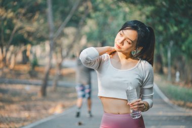 Egzersiz seansından sonra serinleyen düşünceli bir kadın, elinde su şişesi tutarken nazikçe boynuna dokunuyor, ağaçların arasından süzülen yumuşak güneş ışığıyla sakin bir parkın arka planına kuruluyor..