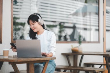 Odaklanmış genç bir kadın, ahşap bir masada birden fazla iş yapıyor. Laptop ve akıllı telefonu arasında rahatça geçiş yapıyor. Yanında da bir fincan kahve..