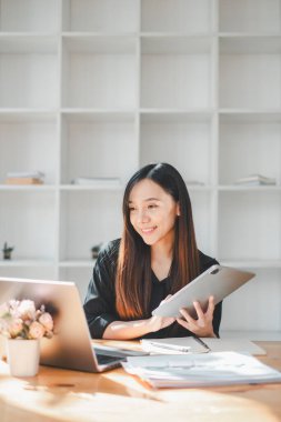 Gülümseyen profesyonel bir kadın dizüstü bilgisayarının yanında dijital tablet kullanır, modern, ışık dolu bir çalışma alanında teknolojiyi kucaklar, dinamik ve bağlantılı bir çalışma tarzının göstergesidir..