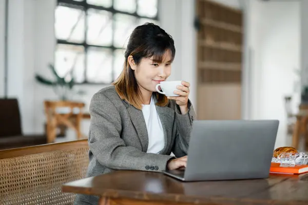 A businesswoman sipping coffee while working on her laptop in a bright, contemporary office setting.