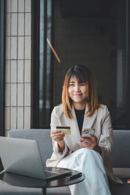 Akıllı telefonuyla meşgul olan modern bir tüketici, elinde bir kredi kartı bulundurur. Finansal veya çevrimiçi işlemlerini yönetmeye hazırdır. Masasında üretkenlik ve çoklu görevi gösteren bir dizüstü bilgisayarla..