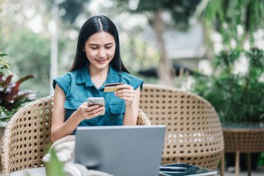 Akıllı telefonuyla online alışveriş yapmak için kredi kartı kullanan, dışarıda dizüstü bilgisayarıyla oturan genç bir kadın..