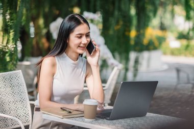 Gülümseyen genç bir kadın dışarıda bir masada dizüstü bilgisayarıyla otururken cep telefonuyla hoş bir sohbet ediyor..