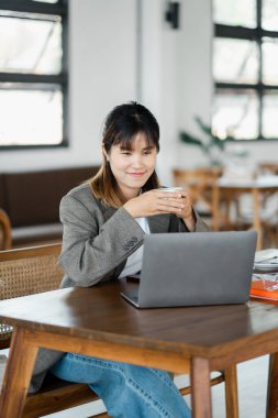 İş kadını laptopuyla uğraşırken masasında hoş bir gülümsemeyle bir fincan sıcak kahvenin keyfini çıkarıyor..