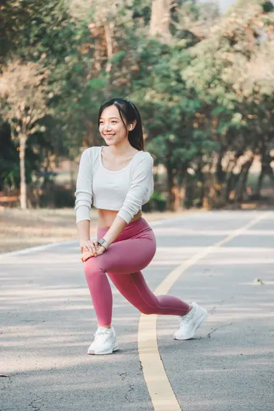 A smiling woman takes a moment to stretch her legs on a running path, wearing pink leggings and white sneakers, with a backdrop of tranquil trees.