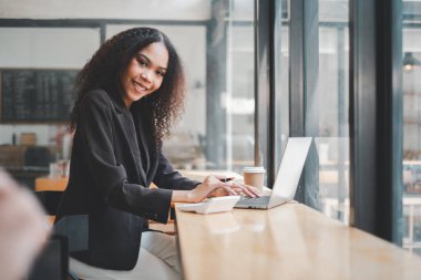 Güneşli bir kahve dükkanında çalışırken gülümseyen kendine güvenen genç girişimci başarılı bir uzaktan çalışma örneği gösteriyor..