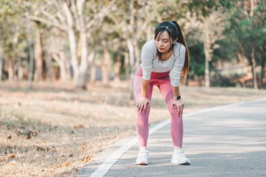 Yorgun kadın koşucu, sakin bir park ortamında koşu yaptıktan sonra nefes almak için ara veriyor..
