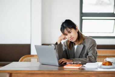 Düşünceli genç profesyonel bir kadın, düşünceli bir ifadeyle ahşap bir masada dizüstü bilgisayarına konsantre oluyor..