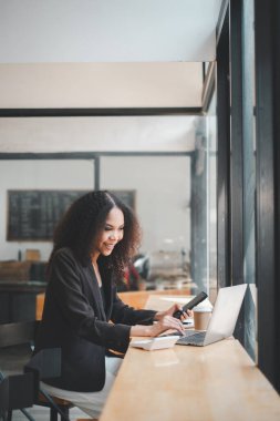 Neşeli genç bir kadın, üretkenlik ve memnuniyet saçan parlak, modern bir kahve dükkanında dizüstü bilgisayarıyla çalışıyor..