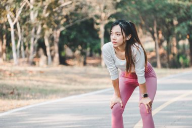 Odaklanmış genç bir kadın koşudan sonra nefesini topluyor, elleri dizlerinin üzerinde, sakin bir park manzarasının önünde, egzersiz sonrası iyileşmeyi vurguluyor..