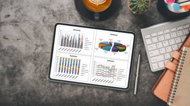 Gri masadaki tablet, kahve, klavye ve kırtasiye malzemelerinin yanında iş özeti grafiklerini gösteriyor..
