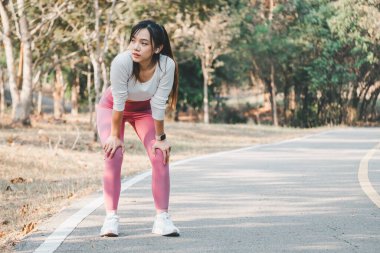 Odaklanmış genç bir kadın koşudan sonra nefesini topluyor, elleri dizlerinin üzerinde, sakin bir park manzarasının önünde, egzersiz sonrası iyileşmeyi vurguluyor..