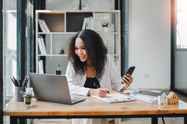 İş kadını, dokümanları incelerken akıllı telefonunu kullanıyor. İyi aydınlatılmış modern bir ofiste, masanın üzerinde bir laptop açık..