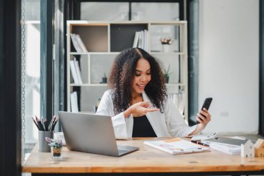 İş kadını, dokümanları incelerken akıllı telefonunu kullanıyor. İyi aydınlatılmış modern bir ofiste, masanın üzerinde bir laptop açık..