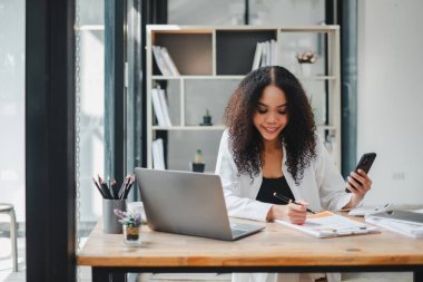 İş kadını, dokümanları incelerken akıllı telefonunu kullanıyor. İyi aydınlatılmış modern bir ofiste, masanın üzerinde bir laptop açık..