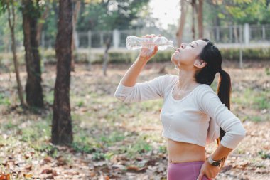 Spor yaptıktan sonra şişeden su içen genç bayan sporcu, doğal park ortamında kendini yeniliyor..