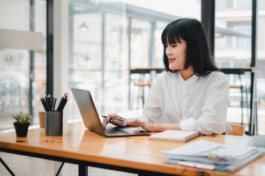 Gülümseyen iş kadını birden fazla görevi, notlar ve raporlarla çevrili geniş bir kafe masasında akıllı telefon ve dizüstü bilgisayar kullanarak.