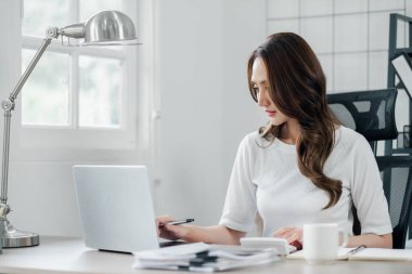 Profesyonel kadın elinde kalemle, elinde dizüstü bilgisayar ve evrak işleriyle masasında çalışıyor..