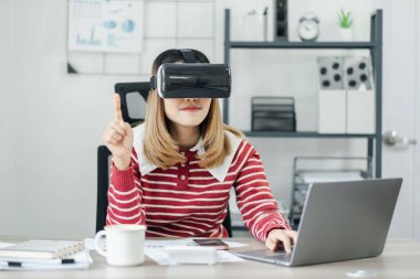 Kırmızı ve beyaz çizgili tişörtlü bir kadın sanal bir ortama giriyor masasında VR kulaklık takarken işaret parmağını kaldırıyor..