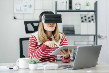 Kırmızı beyaz çizgili süveter giyen kadın ofis masasında sanal gerçeklik kulaklığı takarken tablet kullanıyor..