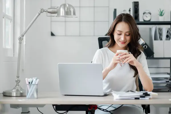 Relaxed professional woman savoring a coffee break at her home office, with a gentle smile and a casual posture, enjoying her workday.