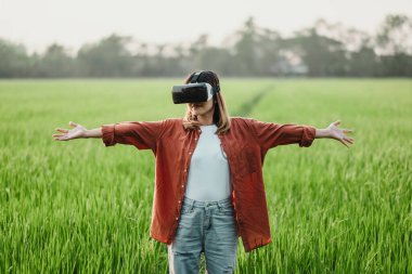 VR kulaklık takan bir kadın çim tarlasında duruyor. Turuncu bir gömlek ve kot pantolon giyiyor.