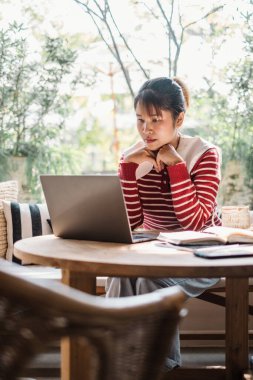 Düşünceli genç bir kadın, parlak bir kafenin yeşillikleriyle çevrili bir anda, laptopundan kafasını çeviriyor..