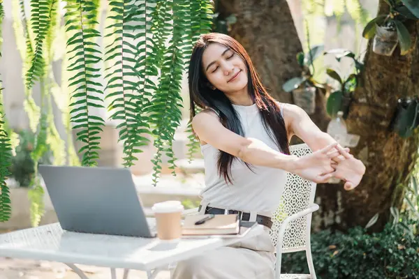 stock image Relaxed female freelancer stretching arms while taking a break from work on her laptop in a lush garden cafe setting