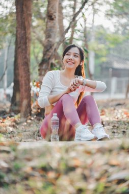 Spor kıyafetleriyle gülümseyen kadın spor yaptıktan sonra spor salonundaki spor izleyicisini kontrol ediyor, sakin bir ormanda oturuyor..