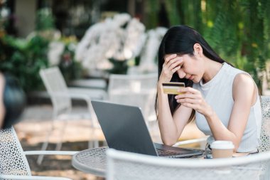 Endişeli genç bir kadın online ödemede sorun yaşıyor, kredi kartı tutuyor ve açık bir kafede dizüstü bilgisayar ekranına bakıyor..