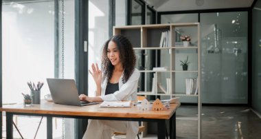 İş kıyafeti giymiş kıvırcık saçlı genç bir kadın iyi aydınlatılmış bir ofiste video konferansı sırasında birini mutlu bir şekilde selamlıyor..