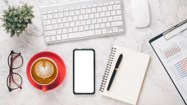 Stylish work desk setup with a keyboard, mouse, smartphone, coffee cup, glasses, and a notebook on a marble surface, ideal for business planning and productivity.