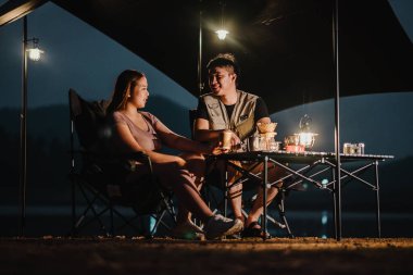Genç çift kamp masasında oturur, sohbet eder ve sıcak bir fener ışığıyla bir çadırın altında dinlenirler..