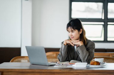 Derin düşüncelere dalmış genç bir kadın masadaki sıcak bir iş yerinde kahve ve kruvasanla dizüstü bilgisayarına bakıyor..