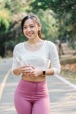 Sıcak gülüşlü, elinde bir su şişesi olan ve dışarıda sakin bir ortamda spor yaparken duraklayan cana yakın genç bir kadın..