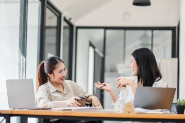 İki nişanlı kadın, bir el hareketi, dizüstü bilgisayarlar ve kağıtlarla parlak bir ofis masasında sohbet ediyorlar..
