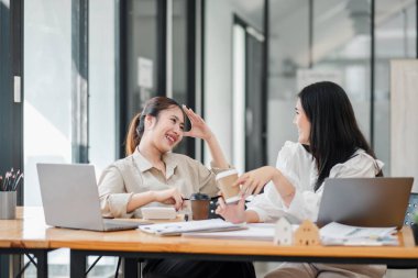 Bir kadın elini alnına koyup gülerken, diğeri elinde bir kahve fincanı tutuyor. İkisi de çalışma masasında dizüstü bilgisayarlarla oturuyor..